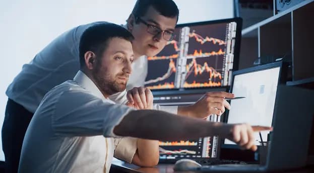 Two people pointing at trading charts on multiple monitors